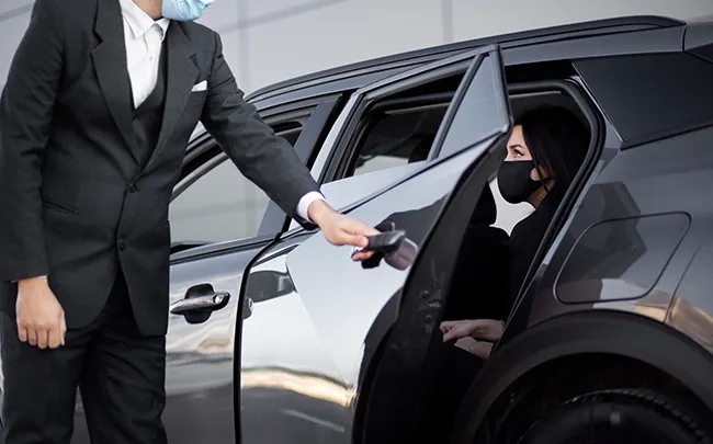 Executive chauffeur welcoming a traveler beside a luxury vehicle for airport pickup.