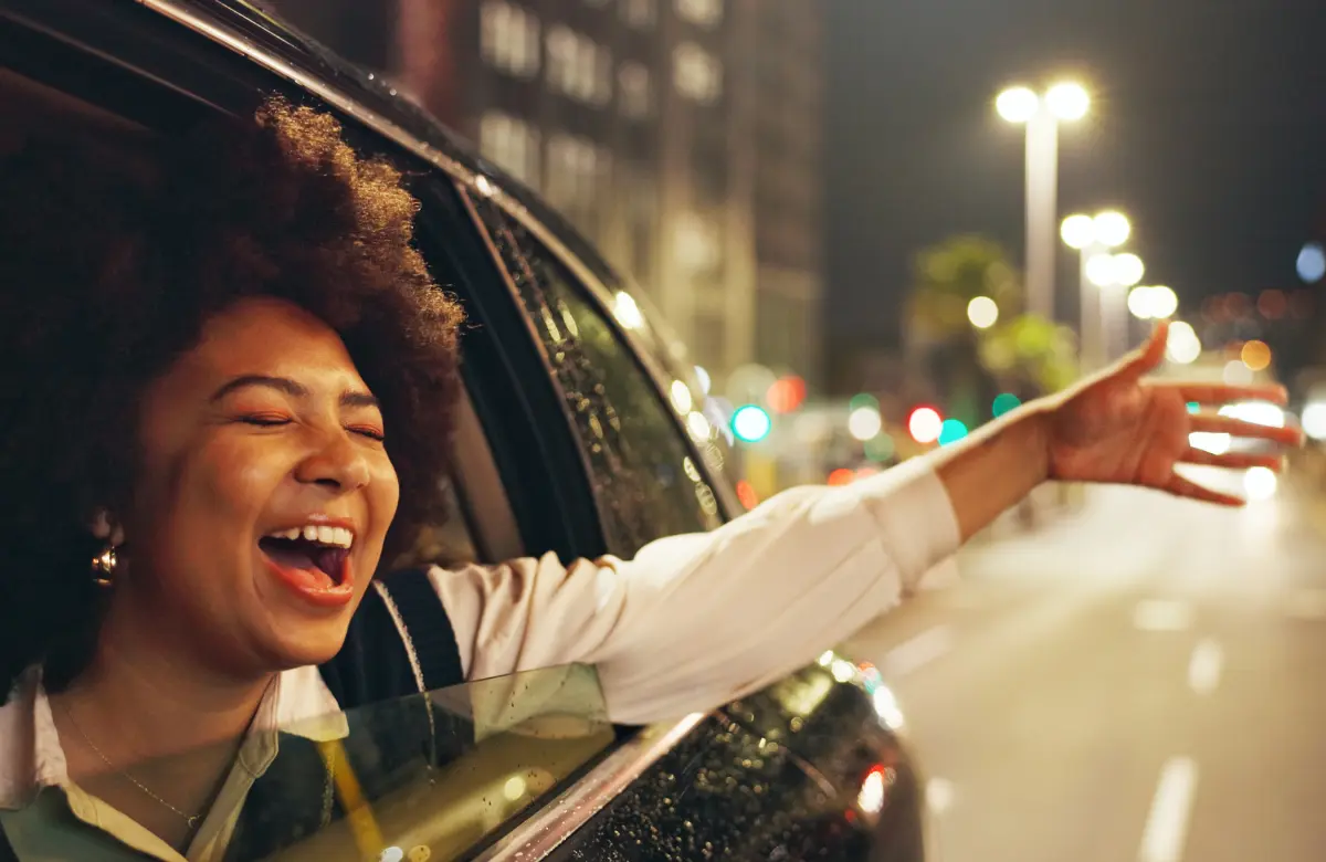 Woman enjoying a late night road trip with her arm out of a car window and city lights in the background.